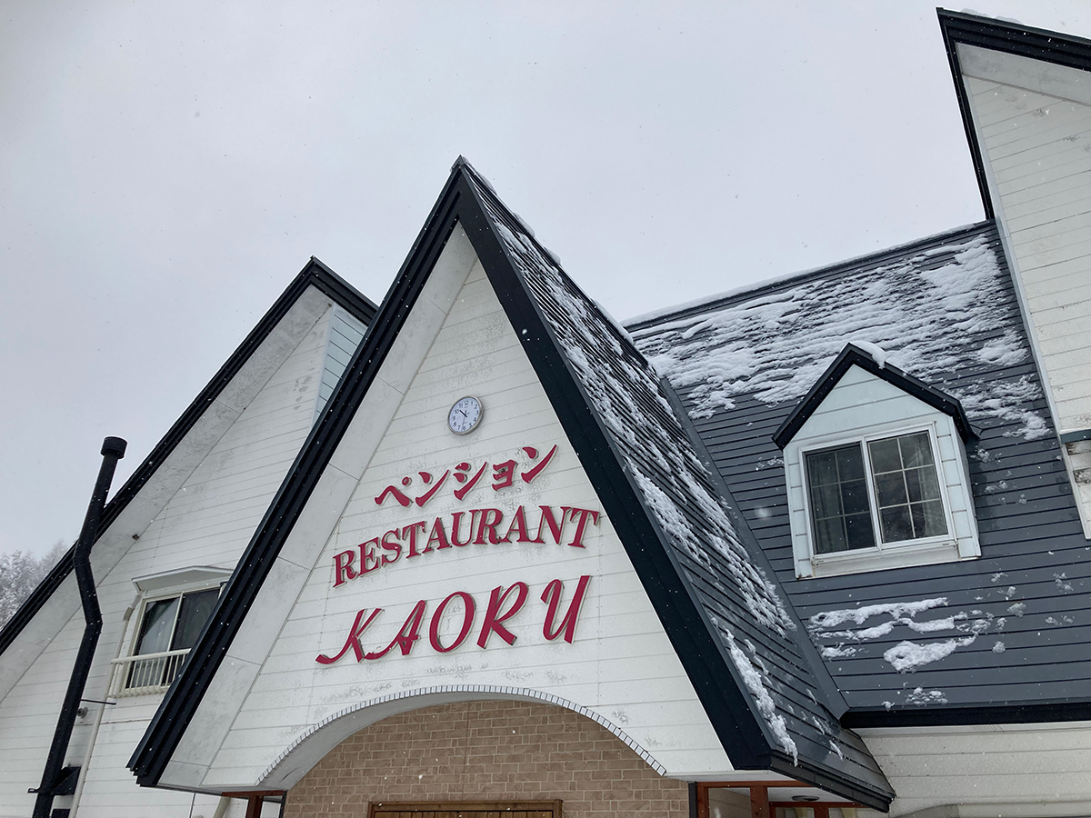 雪が舞う中のペンションかおるの三角屋根と看板