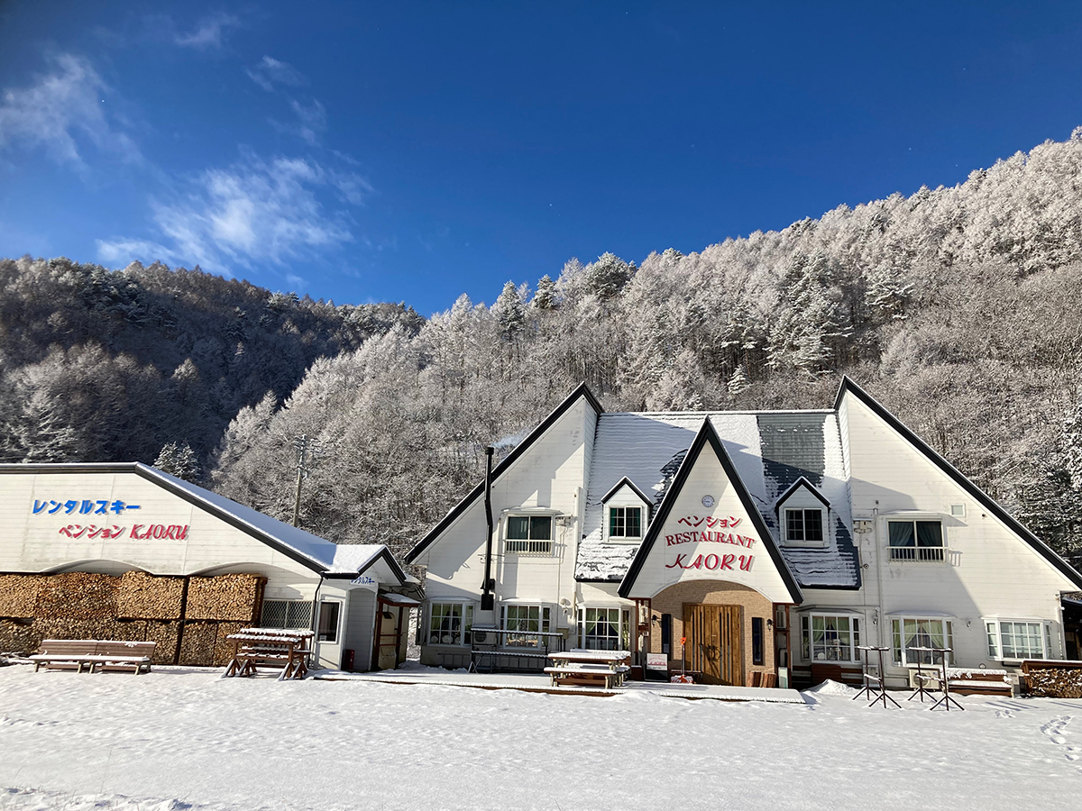 快晴の朝、雪化粧した山を背にしたペンションかおるの外観