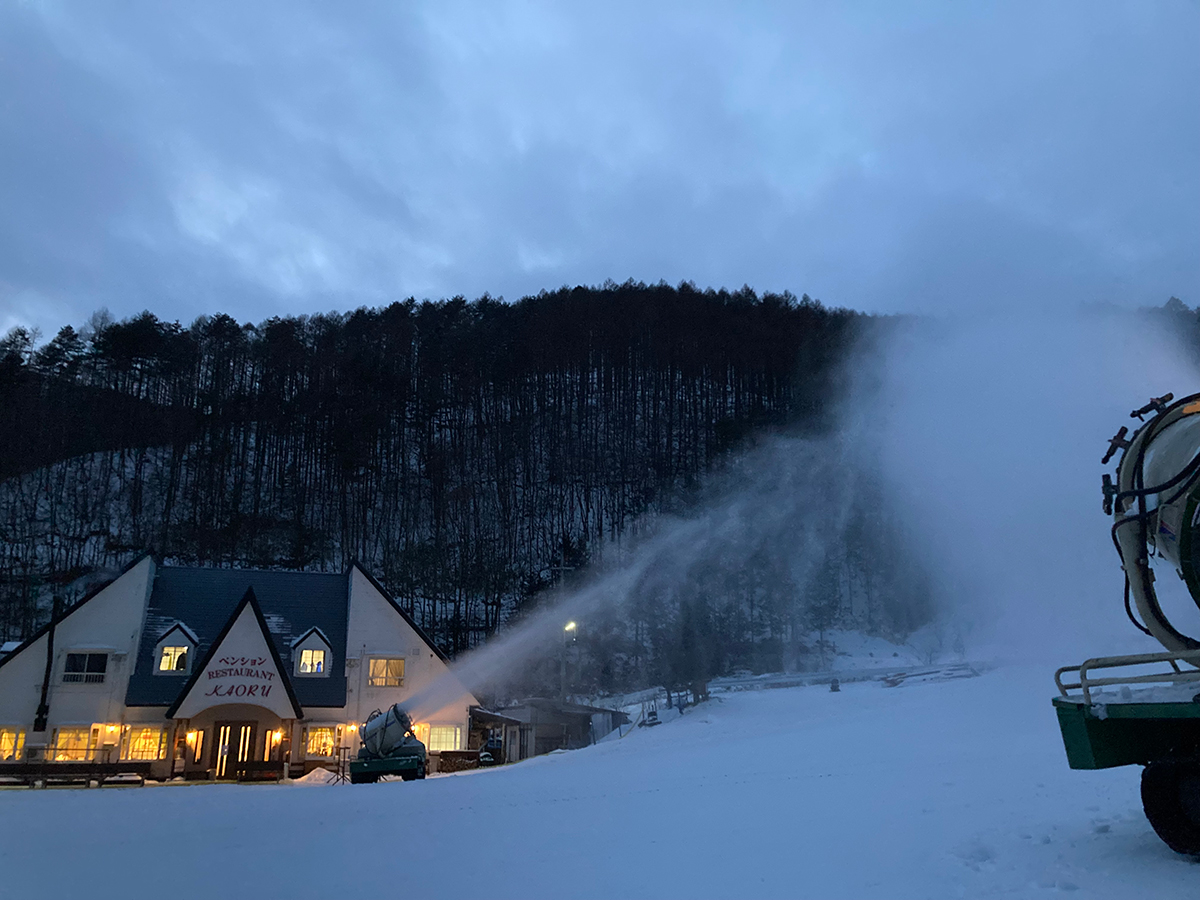夕暮れのゲレンデで人工降雪機が雪をつくる様子と明かりの灯るペンションかおる